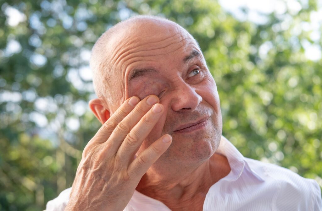 A person standing outside in a park rubs their eye due to dry eye irritation.