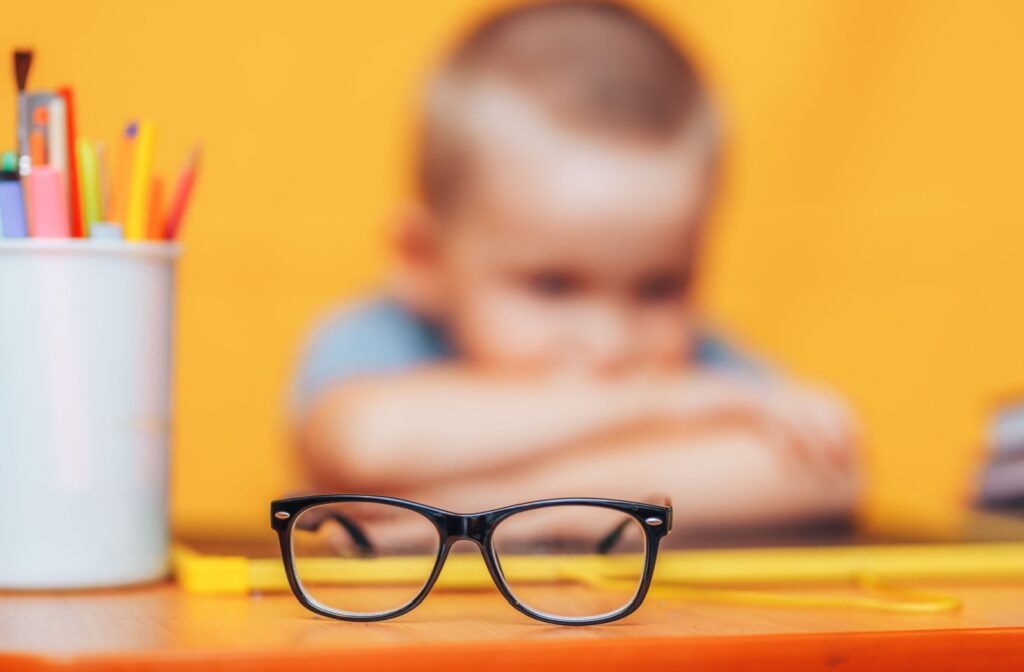 A pair of black glasses on a table with a child blurred in the background, head resting on their arms.